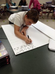 Male student is writing his descriptions to for his Great Lakes project on a large sheet of white paper. 