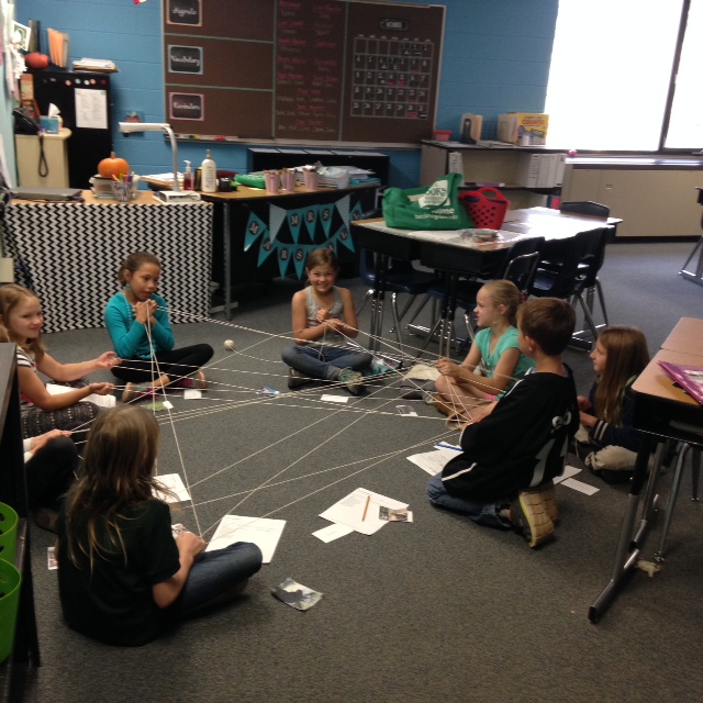 A small group of eight students sits on the floor. These students are in the process of modeling a food web. Each student holds part of one long piece of string that is shared throughout the group based on the interactions that their choose animal would have in the wild. When students tug on the string the other student can feel the interconnections within the ecosystem they are modeling. 